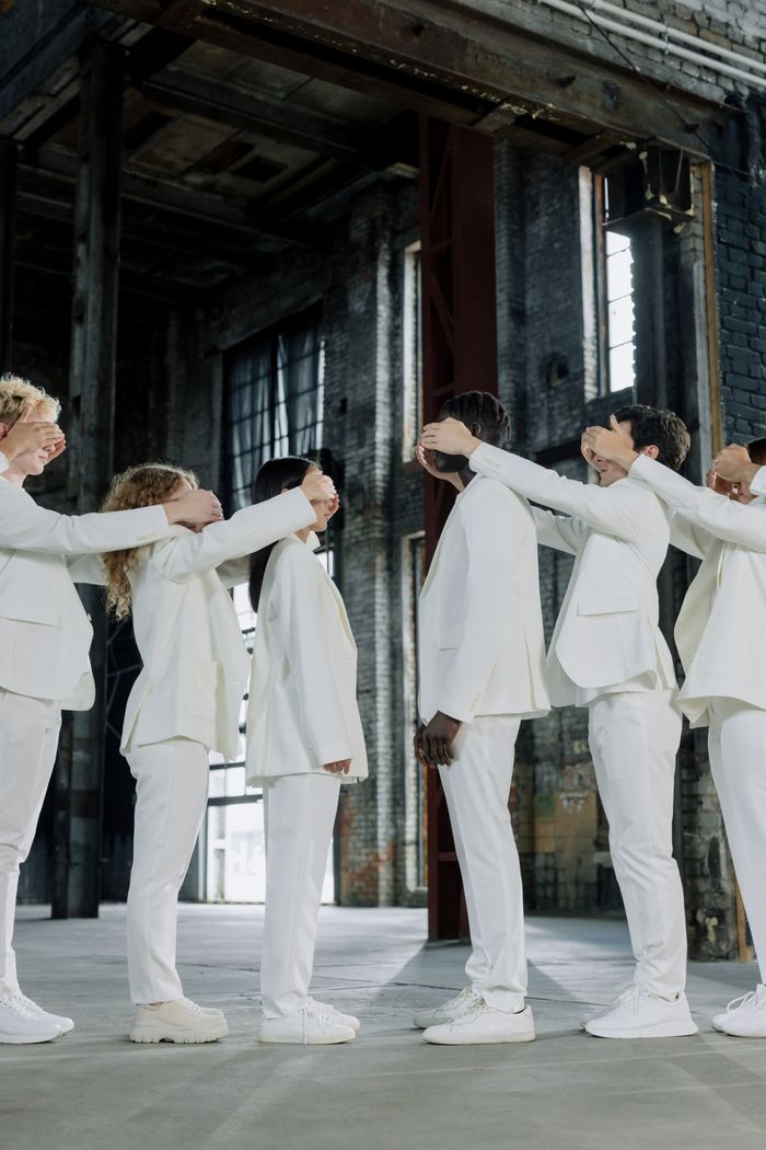 Diverse group in white suits covering each other's eyes in an industrial building.